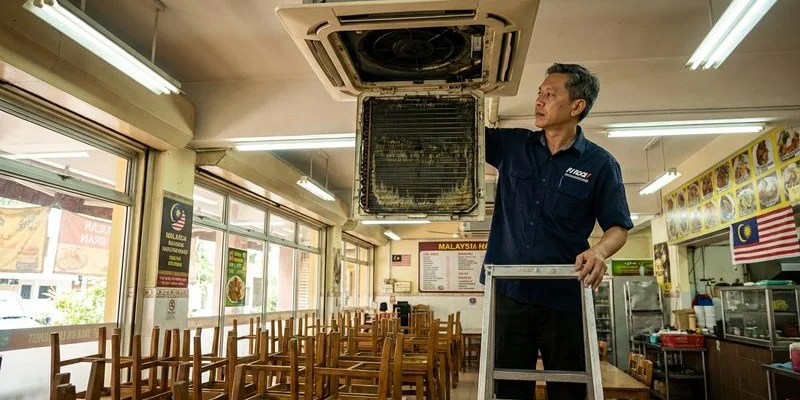Ceiling cassette aircond unit in restaurant dining area being serviced by technician on ladder for monthly maintenance